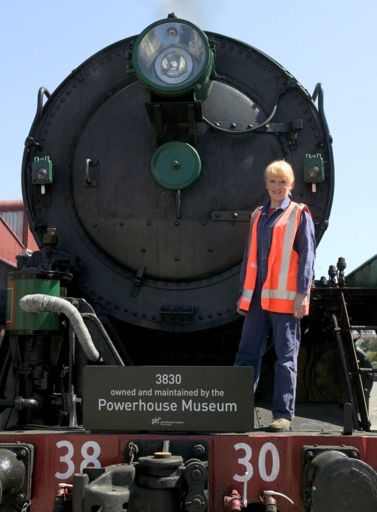 A woman wearing overalls and a 'high-viz' vest is standing on the front of a huge steam locomotive which is in steam. A board on the front of the steam locomotive has the wording "3830 owned and maintained by the Powerhouse Museum". 